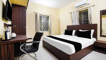 A hotel room with a bed, a desk with a chair, a television, an air conditioning unit, and a window with curtains.
