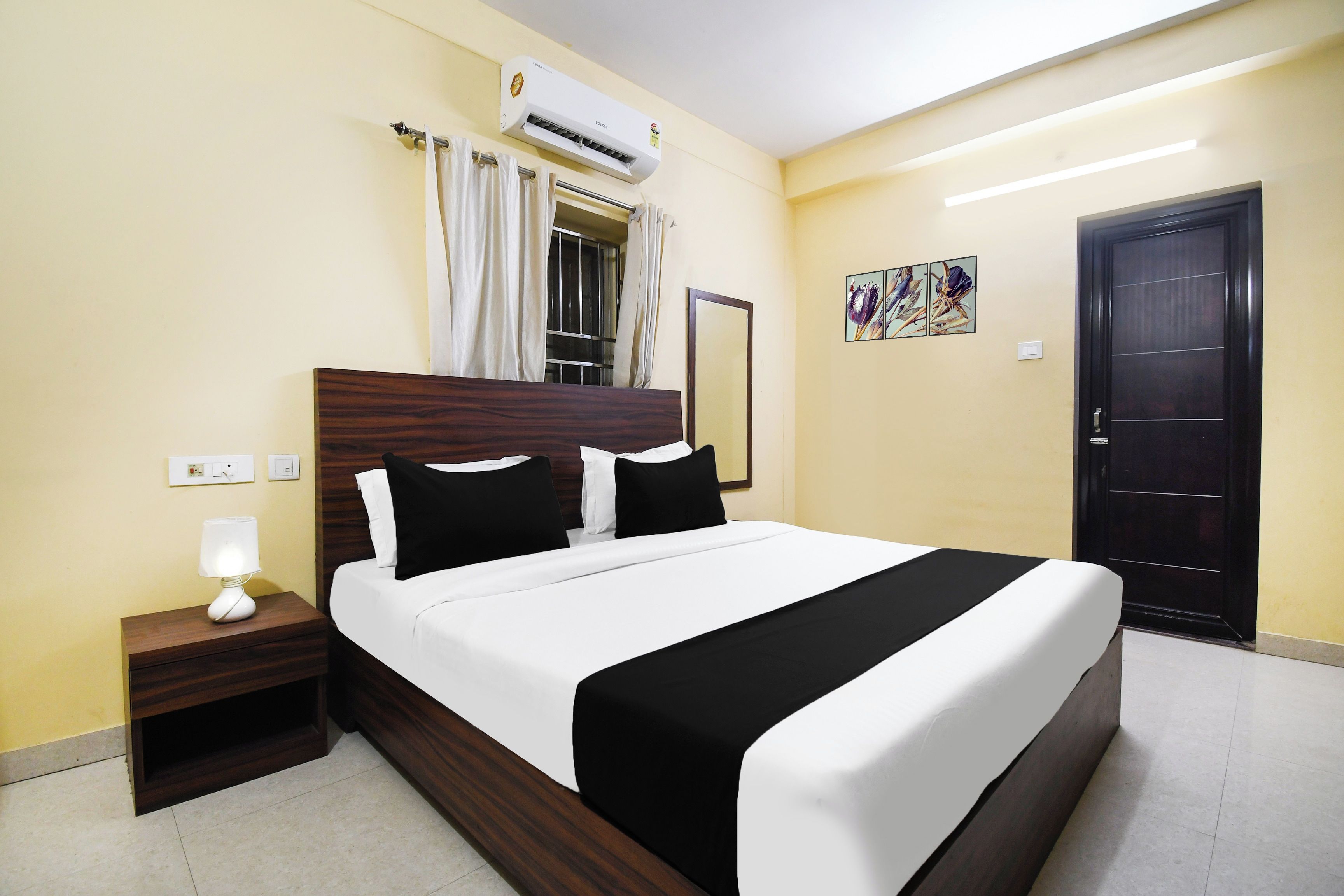 Una camera d'albergo con un letto, un comodino, un condizionatore d'aria, una finestra con tende, una porta e un'opera d'arte appesa al muro.