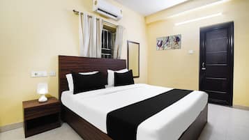 Una camera d'albergo con un letto, un comodino, un condizionatore d'aria, una finestra con tende, una porta e un'opera d'arte appesa al muro.
