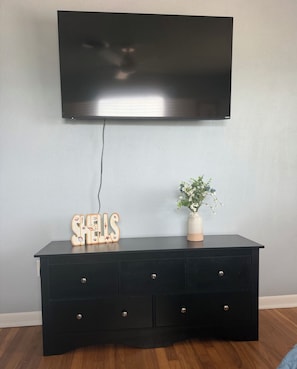 A black TV wall-mounted on a light blue wall, above a black dresser with drawers and a vase with a plant. - Launch Retreat on Merritt Island - Escape to the heart of Florida's Space Coast (Merritt Island)