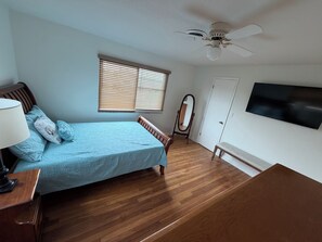 Un dormitorio con cama, cómoda, tocador, ventilador de techo, ventana con persianas y televisión montada en la pared.