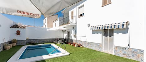 Une piscine dotée d’une terrasse blanche, entourée d’une pelouse bien entretenue, et un bâtiment avec une façade carrelée et un balcon.