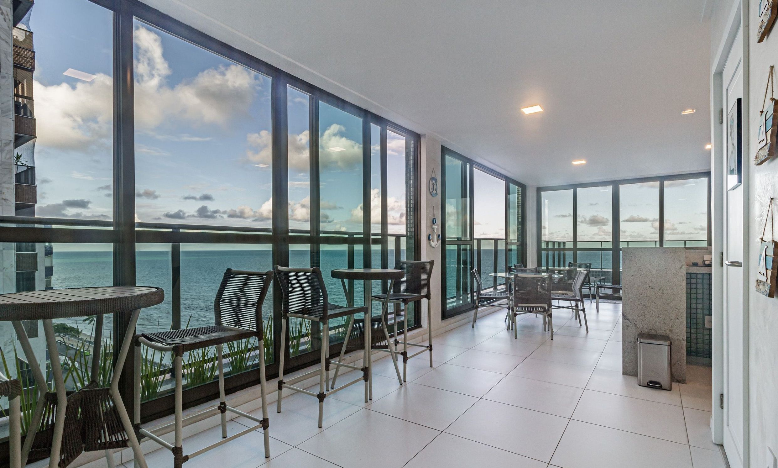 A spacious balcony with large windows overlooking the sea, furnished with chairs and tables.