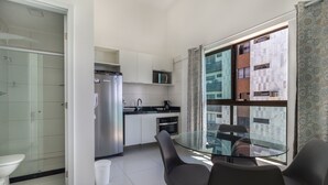 Une cuisine compacte équipée d’un réfrigérateur, d’un four à micro-ondes et d’une cuisinière, une table à manger en verre avec des chaises, et une grande fenêtre donnant sur un immeuble.