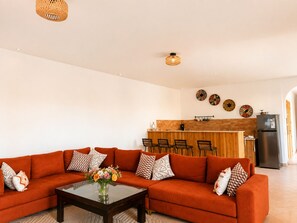 A living room with a sofa, coffee table, and decorative pillows. There is a kitchen area with a counter and chairs in the background. - Villa Tawar - Marrakech (Marrakech)