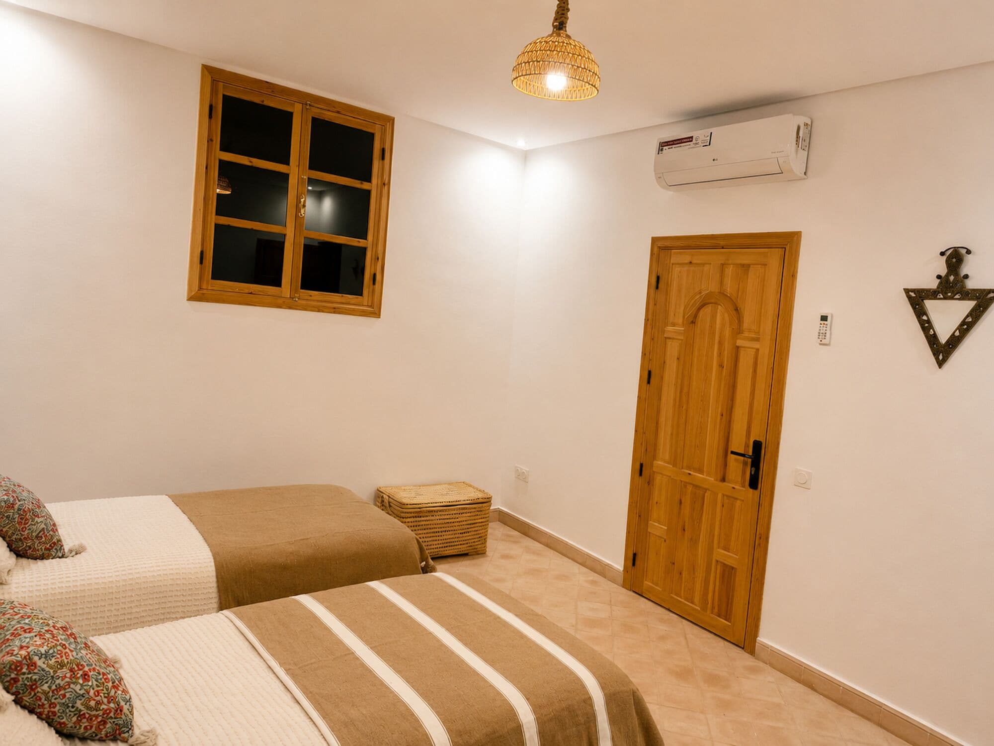 A bedroom with two beds, a window, an air conditioning unit, a door, and a hanging light fixture.