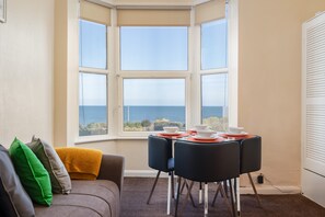A corner room with a bay window overlooking the sea, a dining table with plates and bowls, and a sofa with cushions. - The Blue House  - sleeps 4 guests  in 2 bedrooms (Bridlington)