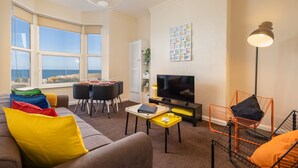 Ein Wohnzimmer mit Sofa, Esstisch, Stühlen, Fernseher und Meerblick durch große Fenster.