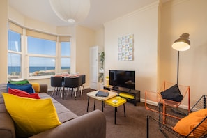 Ein Wohnzimmer mit Sofa, Esstisch, Stühlen, Fernseher und Meerblick durch große Fenster.
