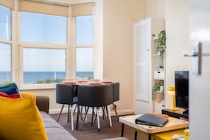 Uma sala de estar moderna com vista para o mar, uma mesa de jantar posta com pratos, e um sofá com almofadas coloridas.