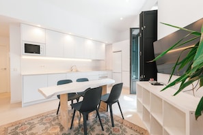 Cozinha moderna com armários brancos, uma mesa de jantar branca com cadeiras pretas e uma televisão grande à direita.