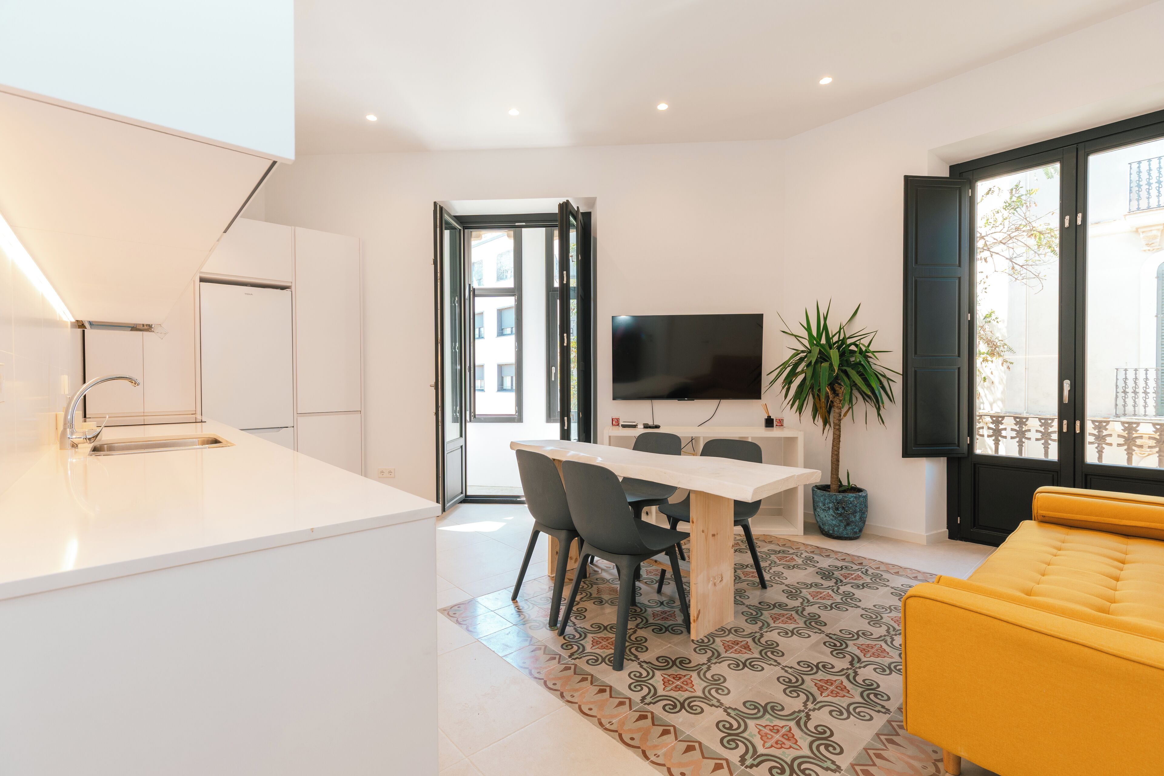 A modern kitchen with a white countertop and sink, a dining area with a table and chairs, a flat-screen TV mounted on the wall, and a yellow sofa.