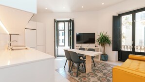 A modern kitchen with a white countertop and sink, a dining area with a table and chairs, a flat-screen TV mounted on the wall, and a yellow sofa.