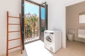A laundry room with a washing machine, a toilet, and a balcony with a view of palm trees and buildings.