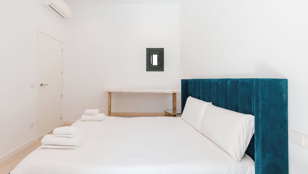 A hotel room with a bed, a bedside table, a wall-mounted air conditioner, and a door.