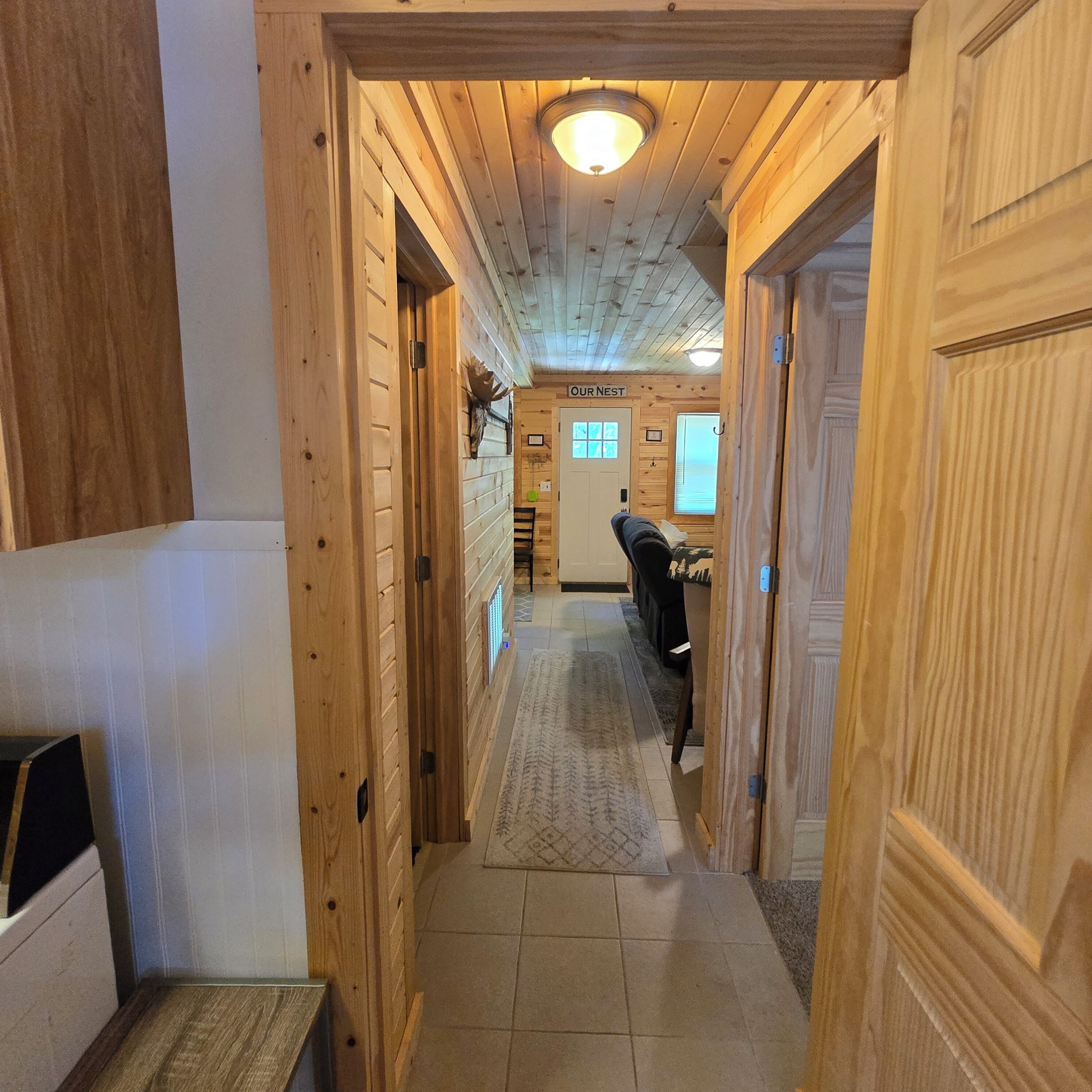 Un pasillo de madera que conduce a una habitación con la inscripción 'OUR NEST', una luz en el techo, una puerta con panel de vidrio y una alfombra en el suelo.
