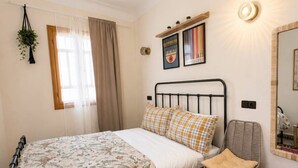 Un dormitorio con cama, una silla, un tapete, una planta colgante y un estante de pared con objetos decorativos.