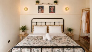 Una cama con colcha estampada, dos mesitas de noche con plantas, una repisa de pared con objetos decorativos y una lámpara colgante.