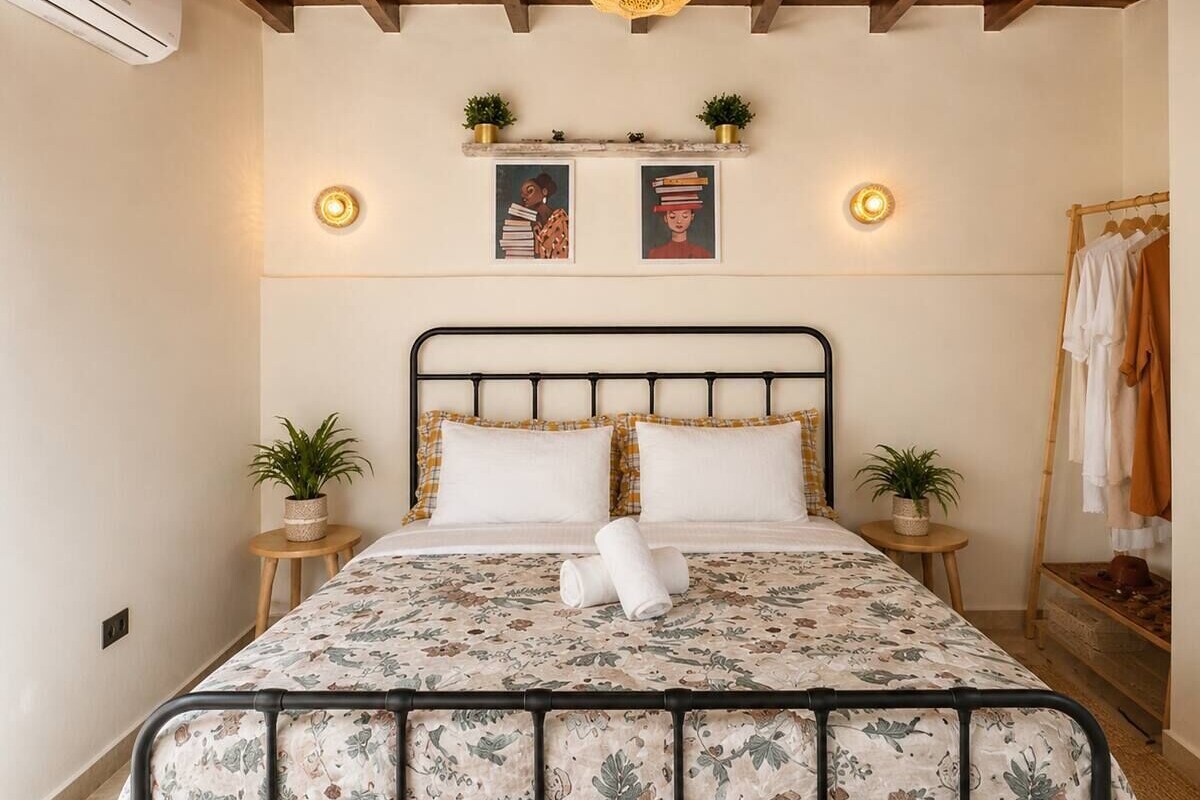 A bed with a floral bedspread, two bedside tables with plants, a wall-mounted shelf with decorative items, and a hanging light fixture.