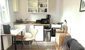 Een kleine keuken met een magnetron, koffiezetapparaat en een prullenbak. Er is een eethoek met een stoel en een tafel.