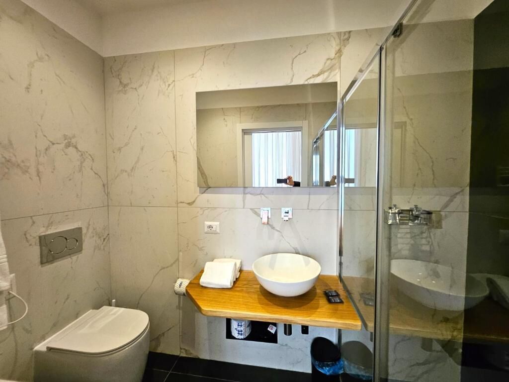 Un bagno moderno con parete in marmo, mobile lavabo in legno, un ampio lavabo bianco, cabina doccia in vetro e un water.