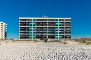 A multi-story hotel with a central atrium, balconies, and a sandy beach in the foreground. - Island Winds East 508 2 Bedroom Condo (Gulf Shores)