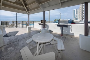 A covered outdoor area with a picnic table and chairs, a barbecue grill, and a view of a pool and buildings in the background. - Island Winds East 508 2 Bedroom Condo (Gulf Shores)