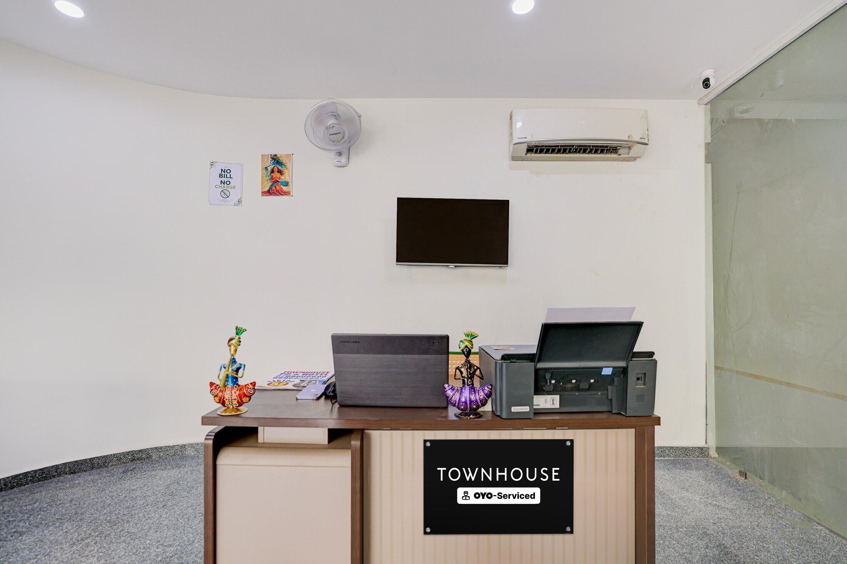 A reception desk with a sign reading 'TOWNHOUSE OYO-Serviced,' a flat-screen TV, a ceiling fan, and a wall-mounted air conditioner.