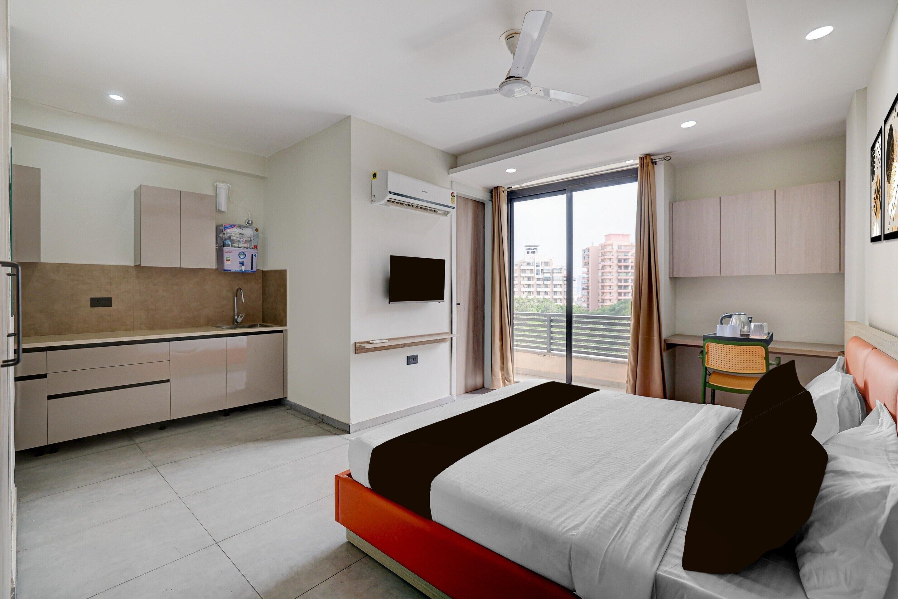 A hotel room with a bed, a TV, a kitchenette, and a balcony with a city view.
