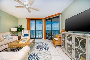 A spacious living room with a large flat-screen TV, a ceiling fan, a sofa, a coffee table, and a view of the ocean through a sliding glass door. - Phoenix X 706 3 Bedroom Condo (Orange Beach)
