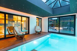 A modern indoor swimming pool with glass walls, wooden flooring, and two lounge chairs.