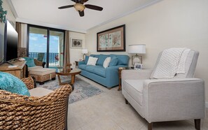 A living room with a blue sofa, a grey armchair, wicker furniture, a ceiling fan, and a view of the ocean. - Phoenix I 1045 2 Bedroom Condo (Orange Beach)