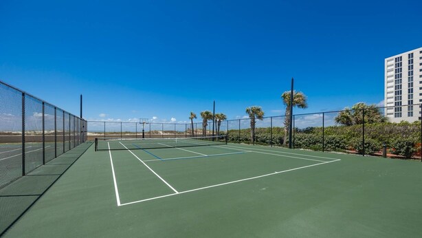 Een tennisbaan met net, omringd door een hek, palmbomen en een gebouw op de achtergrond.