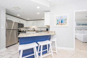 Cocina moderna con armarios blancos, refrigerador de acero inoxidable, encimera de granito y una pared de acento azul. Hay dos taburetes de bar blancos y un cuadro con una escena de playa en la pared.