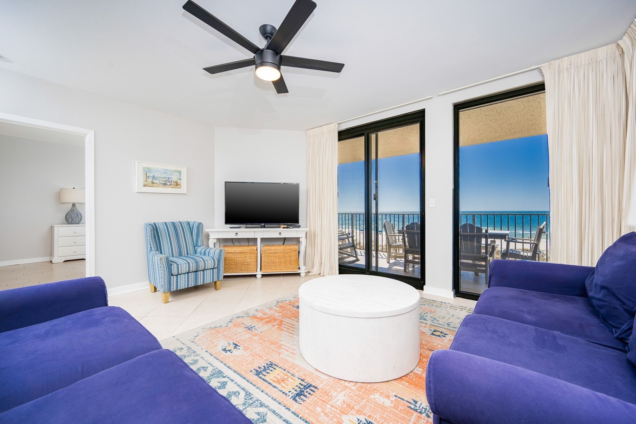 A living room with a flat-screen TV, a ceiling fan, and a view of the ocean through large glass doors.