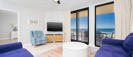 A living room with a flat-screen TV, a ceiling fan, and a view of the ocean through large glass doors.