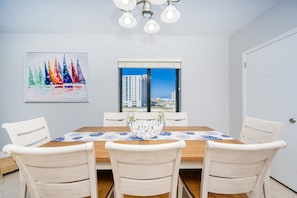 A dining area with a wooden table, white chairs, a white vase, and a painting of sailboats. - Phoenix X 401 3 Bedroom Condo (Orange Beach)