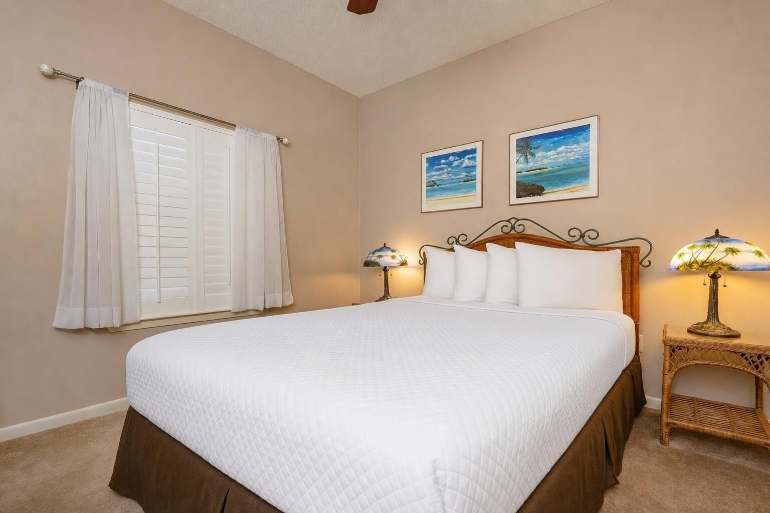A bedroom with a large bed, a window with blinds, two wall-mounted pictures, and a bedside lamp.