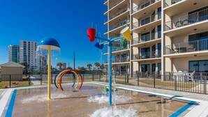 Un espace de jeux aquatiques doté de nombreuses fontaines et d’anneaux colorés, entouré d’une piscine clôturée et d’un immeuble à plusieurs étages avec des balcons.
