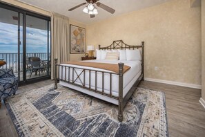 Ein Schlafzimmer mit einem Bett, einem Deckenventilator, einem großen Fenster mit Vorhängen, Blick auf den Strand und einem gemusterten Teppich.