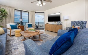 A modern living room with a large flat-screen TV, comfortable seating, a wooden coffee table, and a view through large windows. - Phoenix X 307 3 Bedroom Condo (Orange Beach)