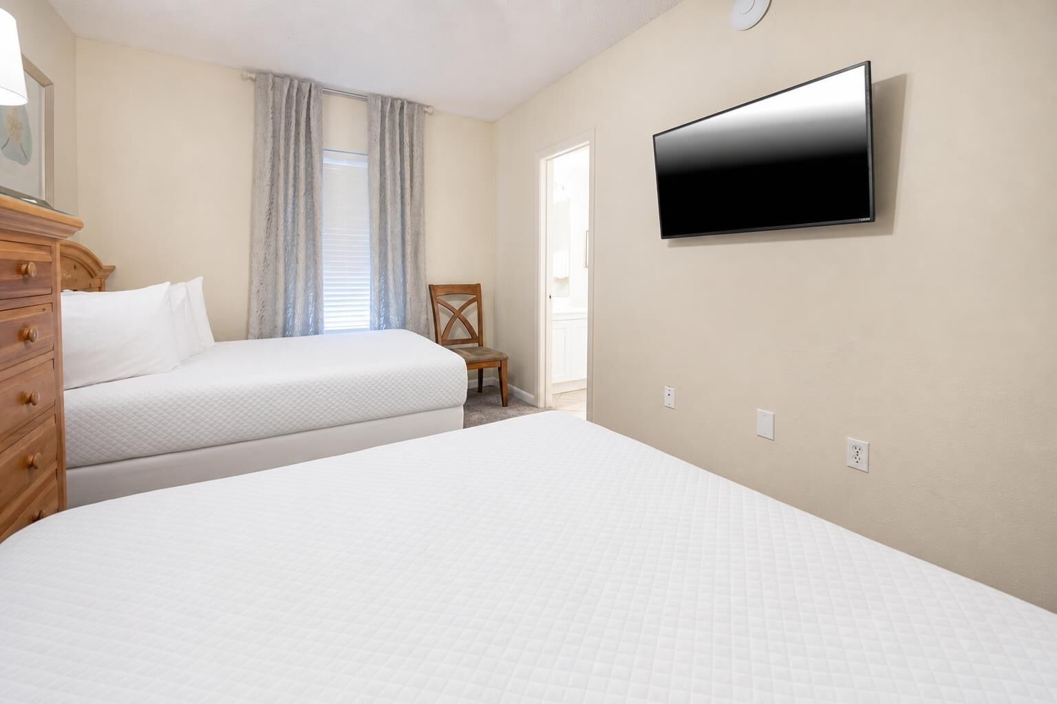 A hotel room with two beds, a wooden dresser, a television mounted on the wall, and a window with curtains.