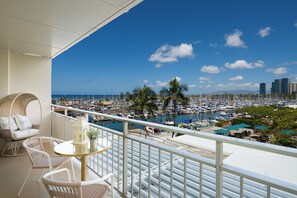 A balcony with wicker furniture, a small table, and a view of a marina with boats and palm trees. - Ilikai 1BR Stunning Harbor/Sunset/Ocean Views! (Honolulu)
