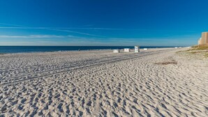 Una extensa playa de arena con huellas que conducen al agua, un cielo azul despejado y algunas personas a lo lejos.