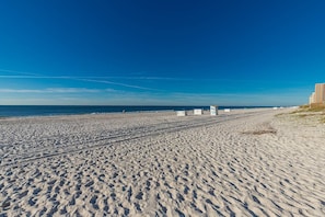 A wide sandy beach with footprints leading to the water, a clear blue sky, and a few people in the distance. - Island Winds East 808 2 Bedroom Condo (Gulf Shores)