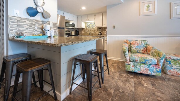 Una cocina con una isla central, taburetes de bar, un sillón estampado y un reloj decorativo en la pared.