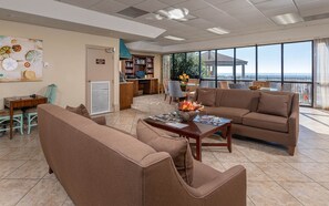 A spacious living area with a large window offering a view of the sea, a comfortable sofa, a coffee table with magazines, and a bookshelf. - Phoenix X 306 3 Bedroom Condo (Orange Beach)