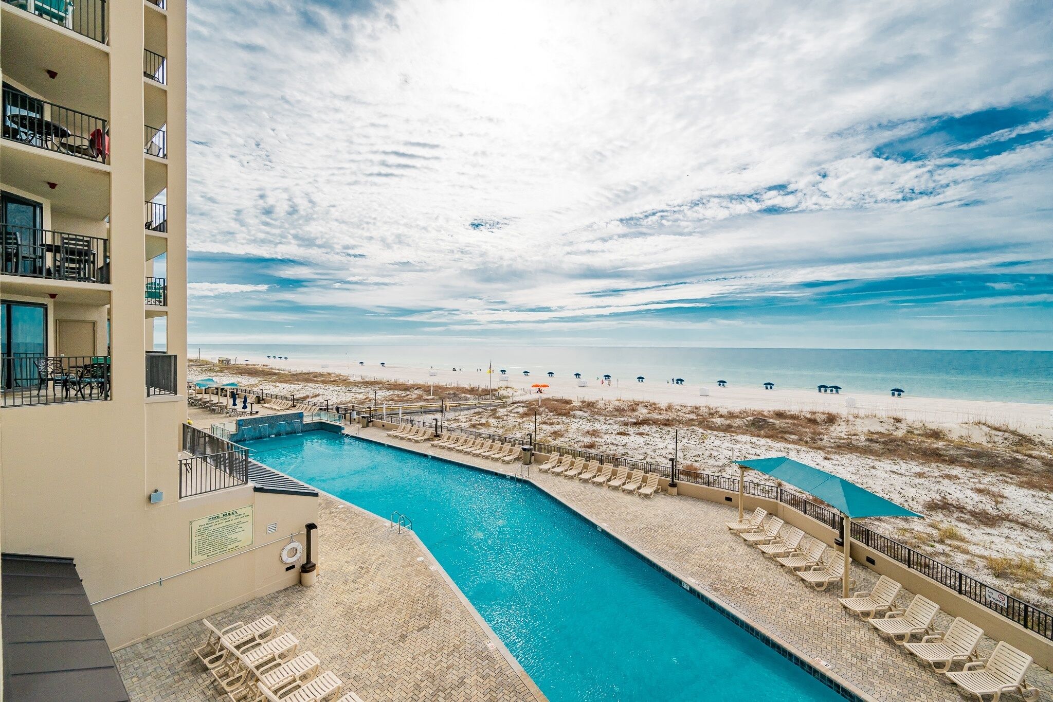 Une piscine avec des chaises longues, une vue sur la plage et un bâtiment doté de balcons.