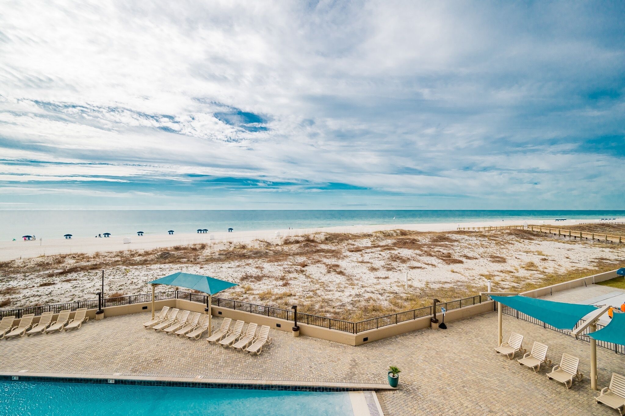 Un área de piscina frente a la playa con sillones de descanso y sombrillas, con vista a una playa arenosa y el océano.