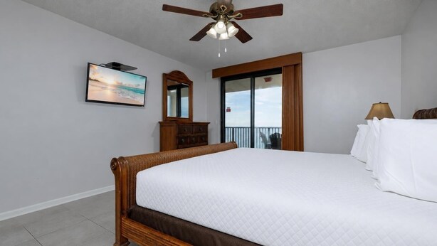 Une chambre à coucher avec un grand lit, un ventilateur de plafond, une fenêtre avec des rideaux, une commode et un tableau encadré au mur.
